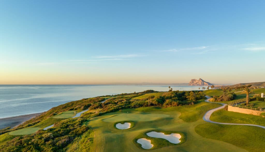 Vista panorámica del campo Links de La Hacienda Golf Resort, con el mar Mediterráneo de fondo y el imponente Peñón de Gibraltar al horizonte. Un paisaje verde y ondulado rodea los hoyos del campo de golf.